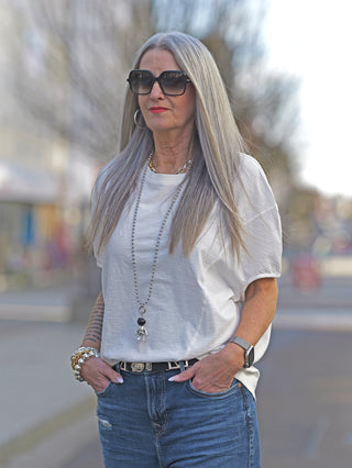 Frau mit langen grauen Haaren trägt das Nuomeno Concept Tee Trapezio tofu: ärmelloses, oversized T-Shirt mit Rundhalsausschnitt, Jeans kombiniert, Sonnenbrille und Kette als modische Akzente.