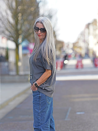 Ein Frau trägt das Nuomeno Concept Tee Trapezio grafite, ein ärmelloses, oversized graues Shirt mit Rundhalsausschnitt, während sie auf der Straße steht, kombiniert mit einer Sonnenbrille.