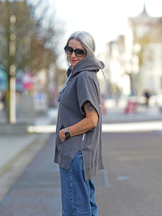 Nuomeno Hoody 14164 Grafite: Frau trägt grauen, ärmellosen Kapuzen-Sweater mit Rückenprint und oversized Passform, kombiniert mit blauen Jeans, Sonnenbrille und Armbanduhr.