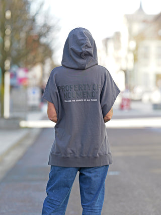 Person trägt den Nuomeno Hoody 14164 Grafite mit Kapuze und Rückenprint. Der ärmellose, oversized Sweater aus 100% Baumwolle wird lässig über einem grauen T-Shirt und blauen Jeans präsentiert.