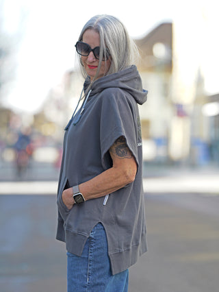 Eine Frau trägt den Nuomeno Hoody 14164 Grafite mit Kapuze und Oversize-Passform. Der graue ärmellose Sweater zeigt ein Rückenprint und wird von einer Sonnenbrille ergänzt.