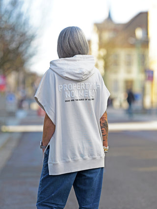 Person trägt den Nuomeno Hoody 14164 tofu mit Kapuze und Rückenprint, oversized Passform. Der Hoodie ist ärmellos und wird mit blauen Jeans kombiniert, auf dem Bürgersteig stehend.