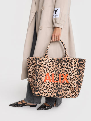 ALIX Leo Shopper, ein Canvas-Shopper mit Leopardenmuster, wird von einer Hand gehalten. Er besitzt einen Magnetknopfverschluss und ein Reißverschlussfach innen. Maße: 34 cm x 58 cm.