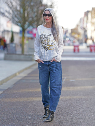 Frau mit Sonnenbrille trägt OFF#DLY Longsleeve Wild Heart. Weißes Shirt mit Herz-Print, kombiniert mit Jeans, steht auf dem Bürgersteig.