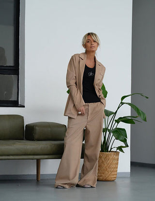 Frau trägt Gitta Banko Hose Camille Sand, eine weite, lässige Hose mit kurzen Bundfalten, stehend vor einem grünen Sofa.