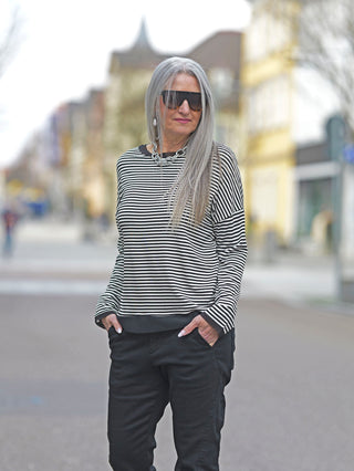 Eine Frau mit langen grauen Haaren und Sonnenbrille trägt das OFF#DLY Ringel Tee Klara in Schwarz/Weiß, ein gestreiftes Rundhals-Longsleeve mit Tüllbündchen, während sie lässig dasteht.