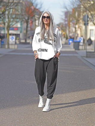 Frau trägt das Rundholz Tee 3290502 mit auffälligem Print, kombiniert mit Sonnenbrille und Kapuzenpullover, beim Spaziergang auf der Straße. Perfekt für künstlerische Street-Fashion.