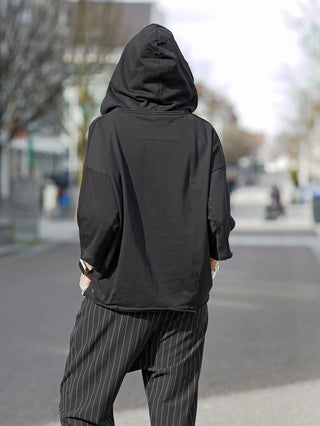 Person trägt das Rundholz Tee 3290502 in Schwarz, kombiniert mit gestreiften Hosen. Das T-Shirt hat einen minimalistischen Stil und eine oversized Passform, ideal für avantgardistische Mode.