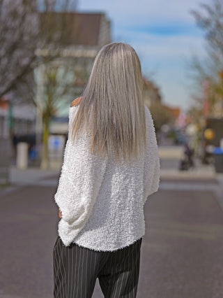 Frau trägt Rundholz Pulli 3480704 offwhite mit Fransen und verkürzten Ärmeln, hinten länger geschnitten. Kombiniert mit gestreifter Hose, im urbanen Umfeld fotografiert.