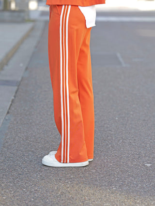 Person trägt OFF#DLY Pant Stripes orange mit fließender, weiter Passform und sportlichen weißen Streifen; kombiniert mit weißen Schuhen für einen dynamischen Look. Elastischer Bund sorgt für Komfort.