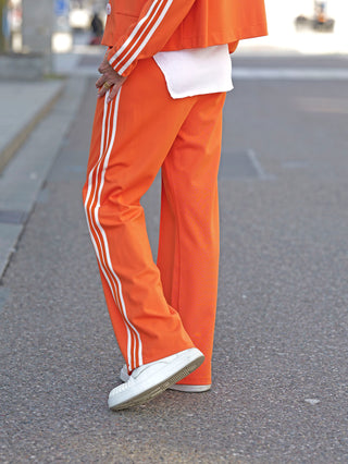 Person trägt OFF#DLY Pant Stripes orange mit lässiger, weiter Passform und weißen Kontraststreifen. Sichtbar sind orangefarbene Hosenbeine und weiße Schuhe auf einer Straße.