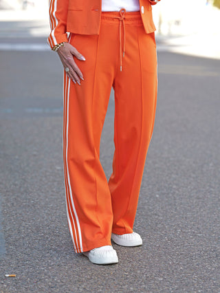 OFF#DLY Pant Stripes orange: Eine Person trägt die orangefarbene Hose mit weißen Streifen, sportlich-lässig mit elastischem Bund und Kordelzug, kombiniert mit weißen Schuhen für einen dynamischen Look.