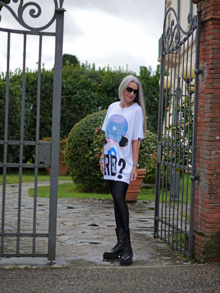Frau trägt OFF#DLY Lon Tee Tina white/blue, ein oversized Shirt mit auffälligem Cartoon-Frontprint, vor einem Metalltor. Kombiniert mit schwarzen Stiefeln für einen urbanen Streetwear-Look.