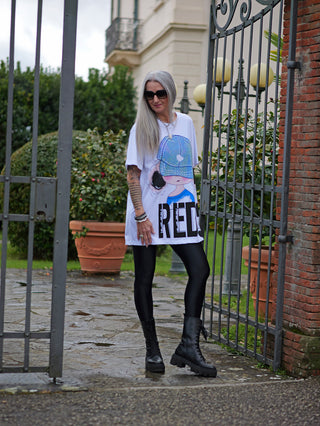 Frau trägt das OFF#DLY Lon Tee Tina white/blue, ein oversized Shirt mit auffälligem Frontprint, stehend vor einem Tor. Kombiniert mit schwarzen Stiefeln für einen urbanen Look.