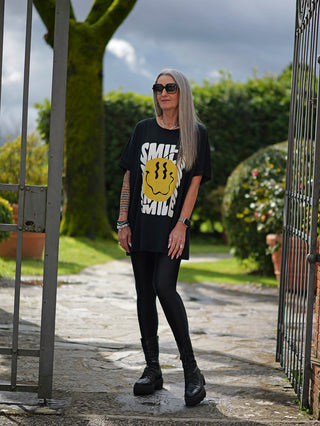 Frau trägt das OFF#DLY Long Tee Crazy Smile, ein oversized Shirt mit großem Smiley-Print, kombiniert mit schwarzen Stiefeln, vor einem Tor stehend. Ideal für lässigen Streetstyle.