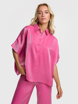 ALIX Shine Boxy Bluse pink: Eine Frau trägt eine glänzende, übergroße Bluse mit kurzen Ärmeln und durchgehendem Knopfverschluss. Das ALIX-Logo ist auf der Rückseite.