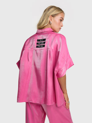 ALIX Shine Boxy Bluse pink: Eine Frau trägt eine übergroße, glänzende pinke Bluse mit kurzen Ärmeln und ALIX-Logo-Bändern. Die Bluse hat einen durchgehenden Knopfverschluss.