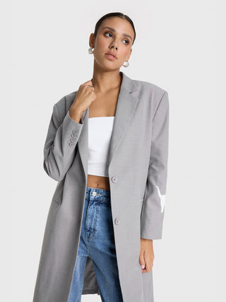 ALIX Clean Long Blazer grey: Frau trägt einen langen, grauen Blazer mit Knopfverschluss, langen Ärmeln, Fronttaschen und einem „ALIX volume 1“-Aufnäher. Perfekt kombiniert mit Polaroid-T-Shirt und Baggy-Jeans.
