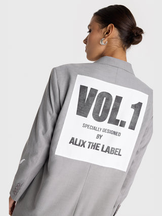 ALIX Clean Long Blazer grey: Frau in langem, grauem Blazer mit weißem Logoaufnäher hinten, übergroßer Passform, Knopfverschluss vorne, langen Ärmeln, Fronttaschen. Kombiniert mit Polaroid-T-Shirt und Baggy-Jeans.