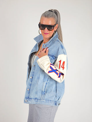 Frau trägt ALIX Mixed Sleeves Denim Jacket denimblue, oversized mit gemischten Ärmeln, sportlichen Patches und „A“-Print auf dem Rücken, kombiniert mit großen Sonnenbrillen für lässigen Streetstyle.