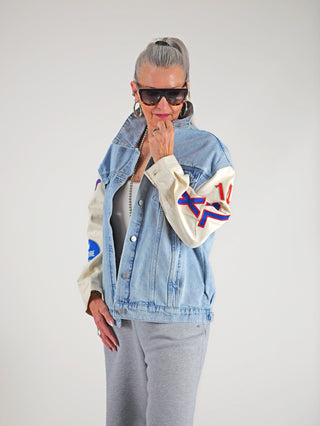 Frau trägt die ALIX Mixed Sleeves Denim Jacket denimblue mit Oversized-Design, Denimkörper und Baumwoll-Ärmeln, dazu sportliche Patches und markante Schriftzüge.