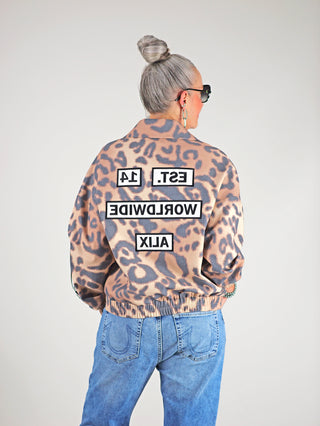 Frau trägt ALIX Jaguar Bomber animal mit Leopardenmuster, doppelreihigem Knopfverschluss, elastischen Bündchen und Logos. Kombiniert mit blauer Jeans, verkörpert der Look urbane Lässigkeit und modischen Streetstyle.