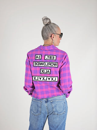 Frau trägt ALIX Cropped Ceck Bluse pink mit Oversized-Passform und ausgefranstem Saum, kombiniert mit einer Jeans. Schriftzug auf Rücken sichtbar. Perfekt für modernen Streetstyle.