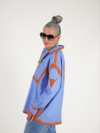 ALIX Sprayed Stripe Bluse softblue: Frau mit Sonnenbrille trägt blau-weiß gestreiftes Oversized-Hemd mit rostfarbenem Spraymuster, klassischem Kragen und Knopfleiste.