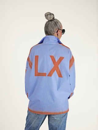 ALIX Sprayed Stripe Bluse softblue, getragen von einer Frau, zeigt ein blau-weiß gestreiftes Design mit markanten rostfarbenen Spray-Effekten, oversized Fit und Statement-Print, ideal für modernen Chic.