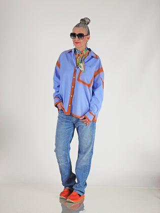 Frau trägt ALIX Sprayed Stripe Bluse softblue, ein oversized blau-weiß gestreiftes Hemd mit rostfarbenen Spray-Effekten, kombiniert mit Sonnenbrille und blauer Jeans für einen lässigen Look.