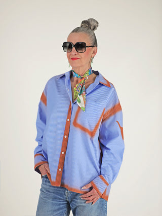 Frau trägt ALIX Sprayed Stripe Bluse softblue mit Oversized-Schnitt und markanten rostfarbenen Spray-Effekten, kombiniert mit Sonnenbrille. Bluse hat klassischen Kragen und Knopfleiste.