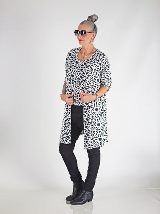 Frau trägt OFF#DLY Leo Set Raelle weiß, bestehend aus lockerem Top und langem Cardigan mit Schwarz-Weiß-Muster, kombiniert mit schwarzen Stiefeln und Sonnenbrille.
