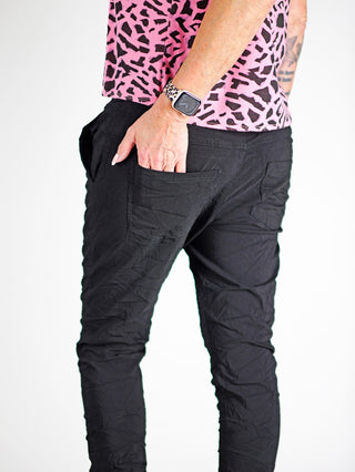 Die OFF#DLY Hose Celeste black zeigt eine schlanke Joggerpant mit Gummibund und Kordelzug, getragen von einer Person mit pinkem Shirt. Eine Armbanduhr ist am Handgelenk sichtbar.