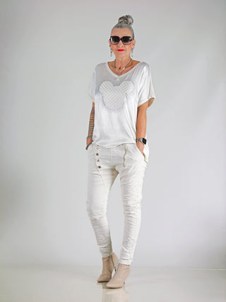 Frau trägt OFF#DLY Shirt Maus vanille mit Glitzer-Print, lockerer Passform und eleganter Materialmix aus Satin und Jersey, kombiniert mit weißen Hosen und Sonnenbrille.