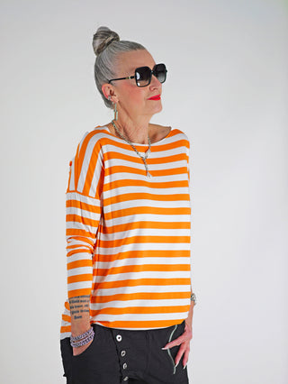 Frau trägt das OFF#DLY Ringel Grosso white/orange, ein gestreiftes Langarmshirt mit Rundhalsausschnitt und langen Ärmeln, kombiniert mit Sonnenbrille und Schmuck, ideal für einen stilvollen, lässigen Look.