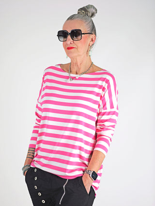 Frau trägt OFF#DLY Ringel Grosso white/pink, ein gestreiftes Langarmshirt mit Rundhalsausschnitt und lockerem Schnitt, kombiniert mit Sonnenbrille für einen stilvollen, lässigen Look.