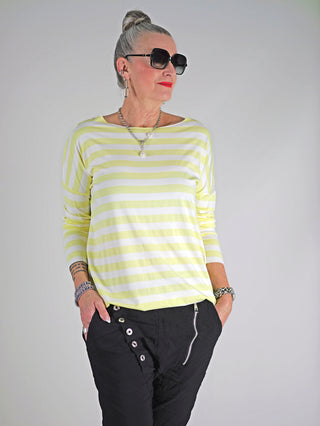 Frau trägt OFF#DLY Ringel Grosso Langarmshirt in Weiß/Gelb mit Sonnenbrille, rotem Lippenstift und Armband. Lockerer Schnitt, Rundhalsausschnitt, lange Ärmel. Perfekt für lässigen, stilvollen Look.