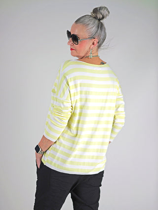 Eine Frau trägt das OFF#DLY Ringel Grosso Langarmshirt in Weiß/Gelb mit Streifen, kombiniert mit einer Sonnenbrille. Das Shirt hat lange Ärmel und einen lockeren Schnitt.