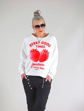 Eine Frau trägt den OFF#DLY Sweat Berry white, ein lässiger Sweater mit Erdbeer-Print und Schriftzug, kombiniert mit Sonnenbrille und weißem Hemd. Perfekt für einen trendigen, entspannten Look.