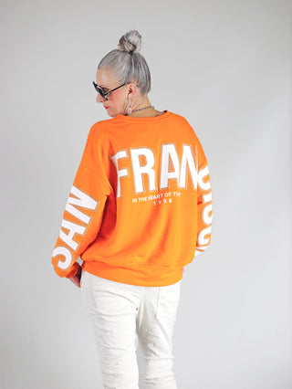 OFF#DLY Sweat San Francisco orange: Frau in oversized Sweatshirt mit „San Francisco“-Schriftzug auf Rücken und Ärmeln, kombiniert mit weißen Hosen. Urbaner Look mit gerippten Bündchen und rundem Ausschnitt.