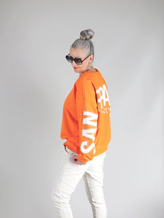 Frau trägt OFF#DLY Sweat San Fransisco orange, oversized mit San Francisco Schriftzug, Sonnenbrille, graue Haare, weiße Hose. Weicher Sweatstoff, Rundhalsausschnitt, gerippte Bündchen, urbaner Streetstyle-Look.
