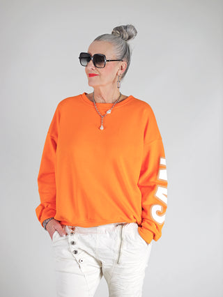 Frau trägt OFF#DLY Sweat San Fransisco orange, Oversize-Sweater mit auffälligem Schriftzug, rundem Ausschnitt und überschnittenen Schultern, kombiniert mit Sonnenbrille und modischem Look.