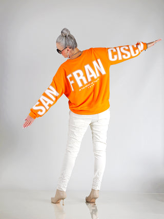 Frau trägt den OFF#DLY Sweat San Francisco orange, einen Oversize-Sweater mit weißem Schriftzug, grauen Haaren und Sonnenbrille, kombiniert mit weißen Hosen und langen Nägeln.