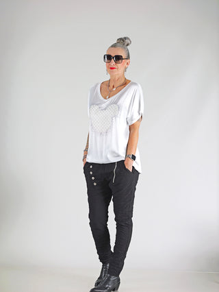 Frau mit Sonnenbrille trägt das OFF#DLY Shirt Maus white mit funkelndem Glitzer-Print, lockerer Passform und angeschnittenen Ärmeln, kombiniert mit schwarzer Hose und schwarzen Stiefeln.