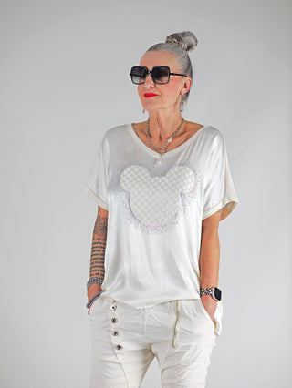 Frau trägt das OFF#DLY Shirt Maus vanille mit lockerem Schnitt, glitzerndem Print und Materialmix, kombiniert mit Sonnenbrille und einer Halskette, für einen stilvollen Alltagslook.