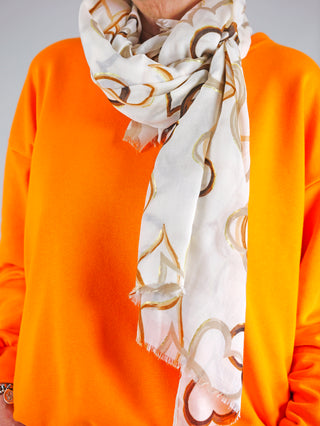 Eine Person trägt den OFF#DLY Schal Heart beige mit goldenen Mustern, kombiniert mit einem orangefarbenen Hemd. Der Schal ist ein modisches Accessoire, das stilvoll um den Hals gewickelt ist.