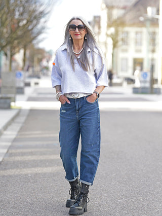 Frau trägt #VDR Polo Sweater grey melange, cropped mit oversized Ärmeln und Kragen-Detail, kombiniert mit Jeans und schwarzen Boots auf dem Gehweg.