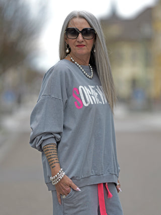 Frau mit Sonnenbrille trägt #VDR Sweat grey, ein graues Oversize-Sweatshirt mit weiß-pinkem Statement-Print SOMEWHERE und pinkem Label am Ärmel, perfekt für lässigen Streetstyle.