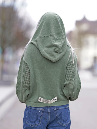 #VDR Pulli olive: Person trägt olivgrünen Strick-Hoodie mit Kapuze, strukturierten Ärmeln und Kängurutasche. Label-Patch auf der Rückseite. Kombiniert mit blauer Jeans für modernen Streetstyle-Look.