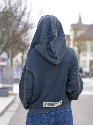 Person trägt den #VDR Pulli black, einen Strick-Hoodie mit Kapuze und Kängurutasche, zeigt strukturiertes Strickmuster an den Ärmeln und ein Label-Patch auf der Rückseite.